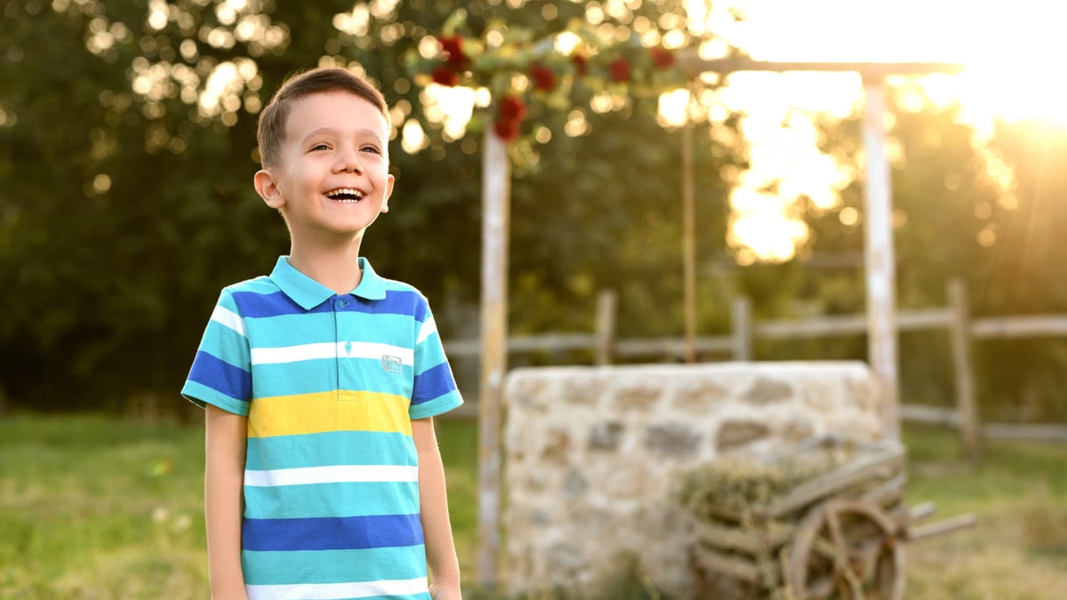 A cheerful boy in a striped shirt smiles joyfully in a sunny garden setting during sunset.
